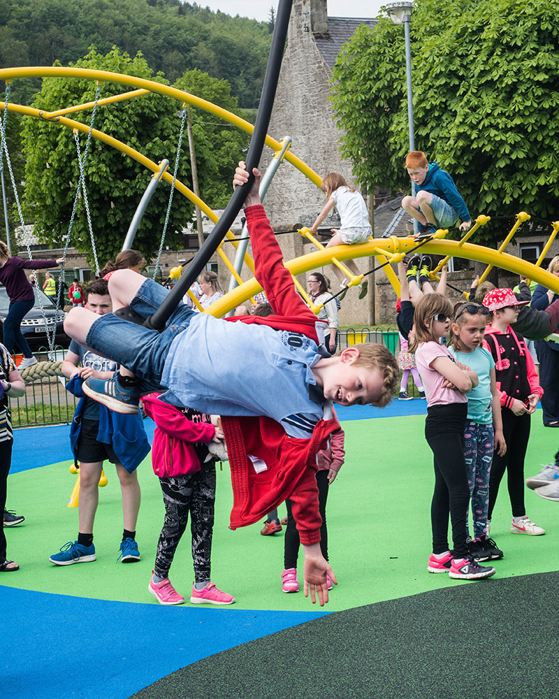 A young boy swings off a zipline seat at a playground which looks very busy in the background.